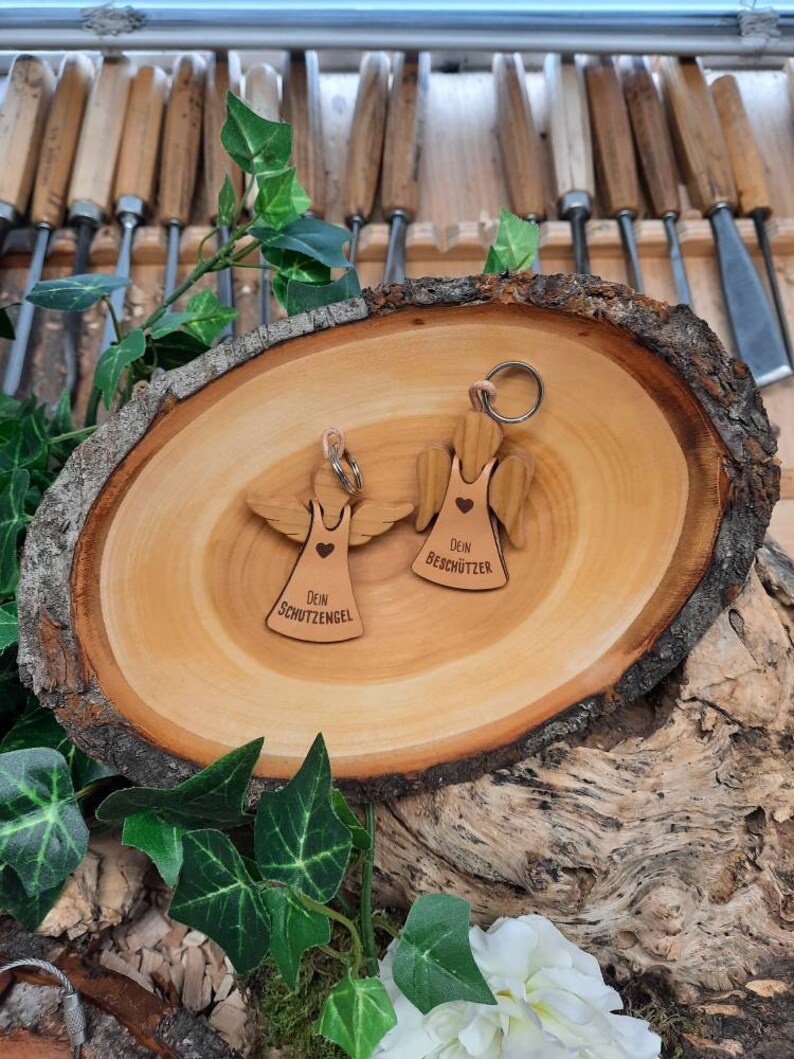 May include: Two wooden angel keychains with the text "Dein Schutzengel" and "Dein Besch&uuml;tzer" on them. The keychains are on a wooden surface.