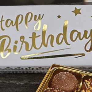 May include: A white box with gold foil lettering that says "Happy Birthday" and gold stars. The box is open and contains a box of assorted chocolates.