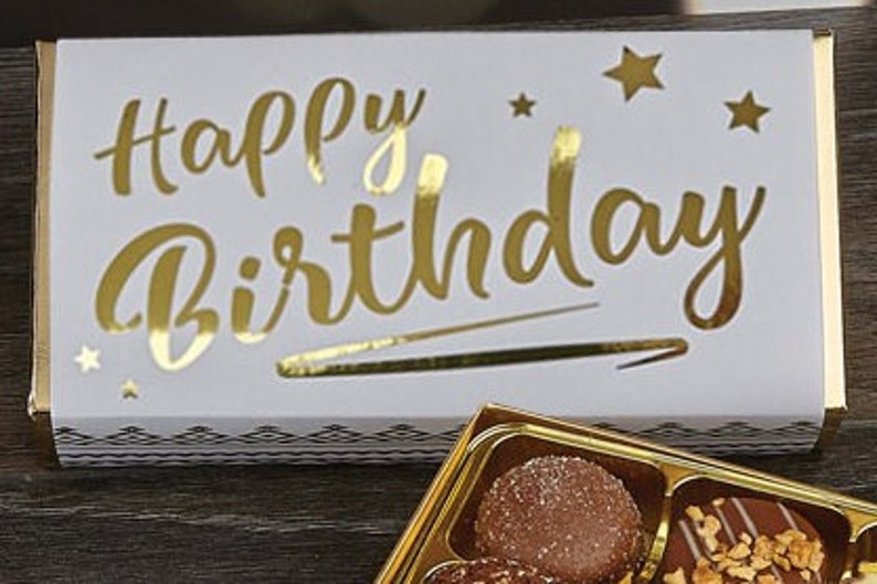 May include: A white box with gold foil lettering that says "Happy Birthday" and gold stars. The box is open and contains a box of assorted chocolates.