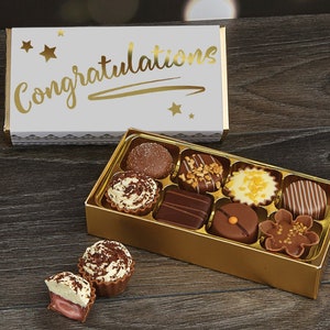 May include: A gold box of assorted chocolates with a white box on top that says "Congratulations" in gold lettering. The chocolates are arranged in a grid pattern and include a variety of flavors and designs.