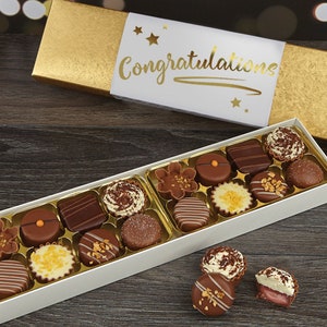 May include: A white box filled with assorted chocolates, with a gold lid that says "Congratulations" in gold lettering. The box is sitting on a wooden surface.