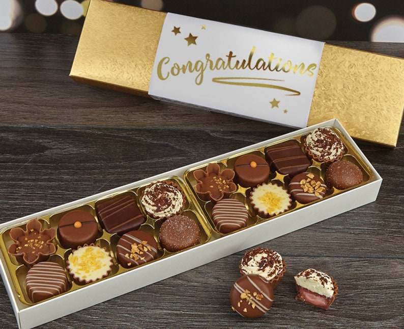 May include: A white box filled with assorted chocolates, with a gold lid that says "Congratulations" in gold lettering. The box is sitting on a wooden surface.