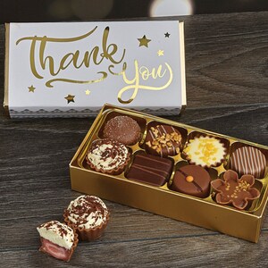 May include: A gold box of assorted chocolates with a white box on top that says "Thank You" in gold lettering. The chocolates are in a variety of shapes and sizes, and some are topped with nuts or sprinkles.