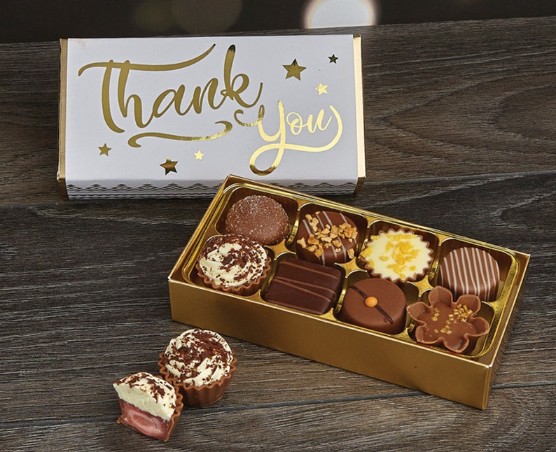 May include: A gold box of assorted chocolates with a white box on top that says "Thank You" in gold lettering. The chocolates are in a variety of shapes and sizes, and some are topped with nuts or sprinkles.