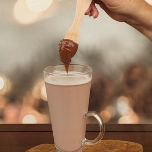 以下が含まれることがあります： 木製のさじにチョコレートヘーゼルナッツスプレッドをつけ、温かいチョコレートドリンクを入れたグラスに浸している様子です