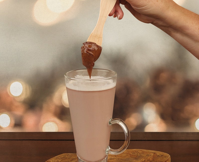 以下が含まれることがあります： 木製のさじにチョコレートヘーゼルナッツスプレッドをつけ、温かいチョコレートドリンクを入れたグラスに浸している様子です