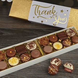 May include: A white box filled with assorted chocolates, with a gold lid that says "Thank You" in gold lettering. The box is sitting on a wooden surface.