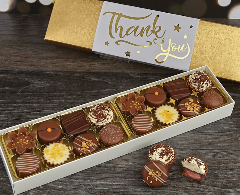 May include: A white box filled with assorted chocolates, with a gold lid that says "Thank You" in gold lettering. The box is sitting on a wooden surface.
