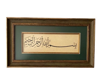 May include: A framed calligraphic artwork with a green mat and a brown wood frame. The artwork features Arabic script in black ink on a cream-colored background. The text reads "Bismillahir Rahmanir Rahim".