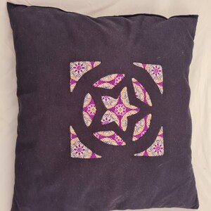 Può includere: Un cuscino nero con un ritaglio a forma di stella in tessuto con motivo viola e oro. Il cuscino è su una superficie bianca.