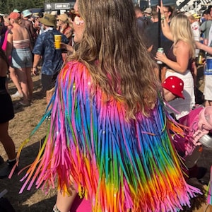 Könnte beinhalten: Eine lebendige, regenbogenfarbene Fransenjacke mit einem Spektrum an Farben, von Pink bis Blau. Die Jacke ist der Fokus, mit langen, fließenden Fransen. Das Bild wurde im Freien auf einem Festival aufgenommen.