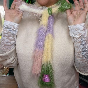 May include: A colorful, fluffy scarf in shades of white, yellow, pink, purple, and green. The scarf has a soft, textured appearance and a label that reads "LOVE Craft". The scarf is held up by a person.