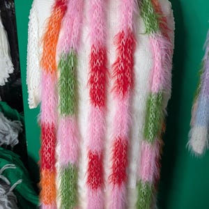 May include: A colorful, fluffy scarf with vertical stripes of pink, red, orange, and green. The scarf is displayed on a white sweater, showcasing its vibrant, textured design. This accessory adds a pop of color.