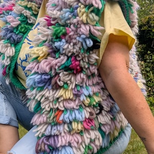 Puede incluir: Un chaleco sin mangas hecho de bucles de lana gruesos y multicolores en tonos rosa, azul, amarillo, verde y blanco. El chaleco presenta una textura densa y peluda creada por la construcción de bucles, sobre una camiseta amarilla clara y pantalones vaqueros azules.