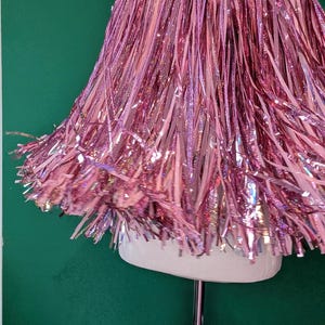 Puede incluir: Una capa confeccionada con numerosas tiras finas de espumillón metálico en tonos rosa y rosa claro. La prenda se exhibe sobre un maniquí, mostrando una silueta acampanada y superpuesta con una textura reflectante y brillante en toda la pieza.