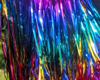 May include: A vibrant rainbow-colored fringe cape, suspended on a black hanger. The cape features long, metallic fringe in a spectrum of colors, including red, orange, yellow, green, blue, and purple. The neckline is adorned with a sparkling trim.
