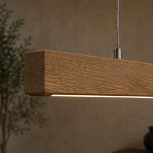 May include: A linear pendant light fitting featuring a rectangular solid wood beam with a visible natural grain pattern. The fitting is suspended by thin metal cables and includes a slim, integrated LED light strip along the bottom edge.