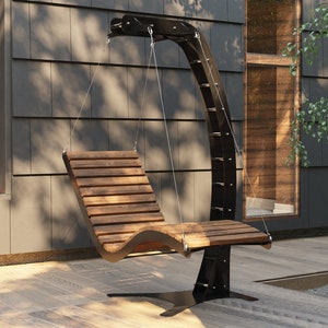 May include: A wooden swing chair hangs from a black metal frame. The chair is made of dark wood slats and has a curved design. The swing is suspended from the frame by metal chains. The swing is in front of a modern house with a gray siding.