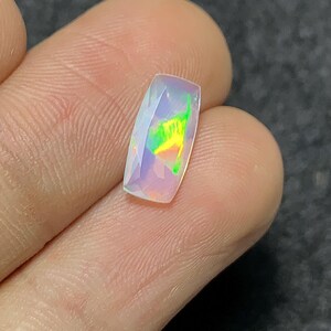May include: A rectangular, faceted opal gemstone with a pale, translucent body. The gem displays internal iridescence with flashes of green, yellow, and orange. The stone is held between fingers, showcasing its polished surface and unique color play.