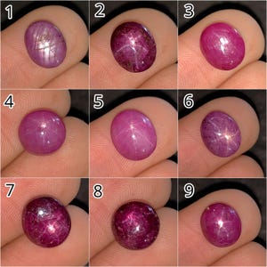 May include: Nine oval and round ruby gemstones in varying shades of red and pink are displayed. Some stones exhibit a star effect, with lines radiating from a central point. The gems are held between fingers, showcasing their polished surfaces.