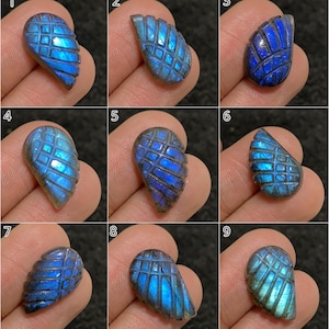 Pode incluir: Nove cabochões de pedras preciosas de labradorita azul com padrões de grade esculpidos. Cada pedra exibe uma forma única e tons variados de azul, com alguns mostrando flashes iridescentes. As pedras preciosas são exibidas em um fundo escuro.
