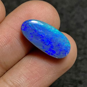 May include: A vibrant blue opal gemstone with a polished, oval shape. The stone displays a play of color with flashes of bright blue and hints of green. The opal is held between fingers, showcasing its size and luster.
