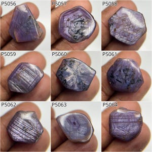 May include: Nine rough gemstones in shades of purple and grey. Each stone has a unique shape and visible internal structures. The gemstones are displayed against a neutral background, highlighting their natural colours and textures.