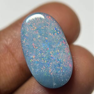 Puede incluir: Gema de &oacute;palo de forma ovalada con un color base azul claro. El &oacute;palo muestra un vibrante juego de colores con destellos de rojo, naranja, amarillo y azul. La gema se sostiene entre los dedos, mostrando su superficie pulida y belleza natural.