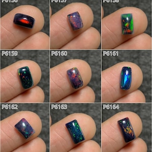 Puede incluir: Nueve piedras preciosas de ópalo pulidas en varios colores, incluyendo negro, azul, verde y rojo. Cada piedra rectangular se sostiene entre los dedos, mostrando el juego de color dentro de los ópalos. Las piedras preciosas están etiquetadas con códigos individuales.