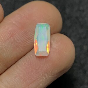 May include: A rectangular opal gemstone with a faceted cut. The opal displays a play of color with flashes of green, orange, and blue. The gemstone is held between fingers, showcasing its brilliance.