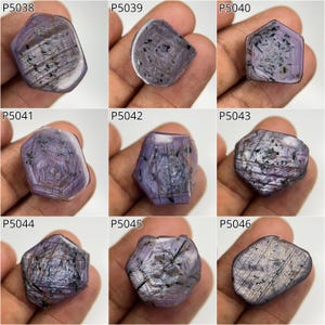 May include: Collection of nine raw, uncut gemstones in shades of purple and gray. Each stone has a unique shape and internal patterns, with visible inclusions and striations. The gemstones are displayed against a neutral background.