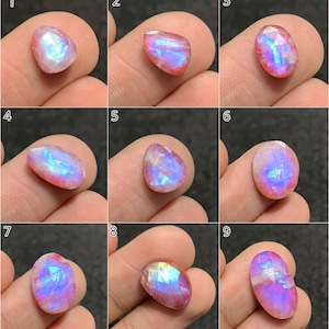 Op de afbeelding: Negen gefacetteerde maansteen edelstenen met een parelmoerglans, in verschillende vormen en maten. Elke steen vertoont een kleurenspel, met tinten roze, blauw en wit. De edelstenen worden tussen de vingers gehouden, waarbij hun individuele kenmerken worden benadrukt.
