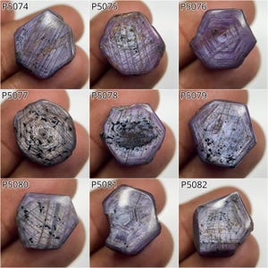 Peut inclure: Neuf pierres précieuses hexagonales brutes, dans des tons violets et gris. Chaque pierre présente un motif unique de lignes et d'inclusions, certaines affichant des zones sombres et mouchetées. Les pierres sont présentées sur un fond neutre.