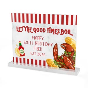 Könnte beinhalten: Ein klarer Acrylschild mit rot-weißen Streifen oben und einem klaren Sockel. Das Schild zeigt den Text „LET THE GOOD TIMES BOIL“ und „HAPPY 60TH BIRTHDAY FRED EST.2026“ mit Illustrationen von Hummer, Mais und scharfer Sauce.