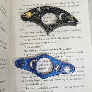 K&ouml;nnte beinhalten: Zwei blaue und schwarze Lesezeichen mit einem Galaxie-Design. Die Lesezeichen zeigen einen Halbmond, einen Stern, eine Rakete und einen Planeten. Der Text "I planned a few public outings to make sure we are at public outings" ist durch die Lupe auf dem blauen Lesezeichen sichtbar.