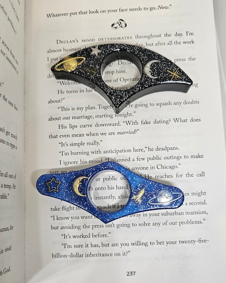 K&ouml;nnte beinhalten: Zwei blaue und schwarze Lesezeichen mit einem Galaxie-Design. Die Lesezeichen zeigen einen Halbmond, einen Stern, eine Rakete und einen Planeten. Der Text "I planned a few public outings to make sure we are at public outings" ist durch die Lupe auf dem blauen Lesezeichen sichtbar.