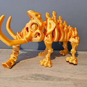 Puede incluir: Un modelo de esqueleto de mamut de plástico de color dorado. El modelo está en posición de pie con las patas separadas y la trompa levantada. El modelo está detallado con costillas, vértebras y huesos de las piernas visibles.