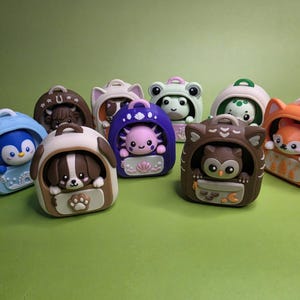 May include: A collection of eight small, colourful plastic animal figurines, each housed inside a matching backpack-shaped carrier. The figures include a penguin, dog, axolotl, owl, fox, frog, and other creatures, each with distinct colours and details.