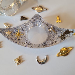 K&ouml;nnte beinhalten: Eine klare, mit Glitzer gef&uuml;llte Ring Schale mit einem Halbmond, Sternen und einem Kometen Design. Die Schale ist umgeben von kleinen goldenen und silbernen Charms, darunter Planeten, Sterne und Raketen.