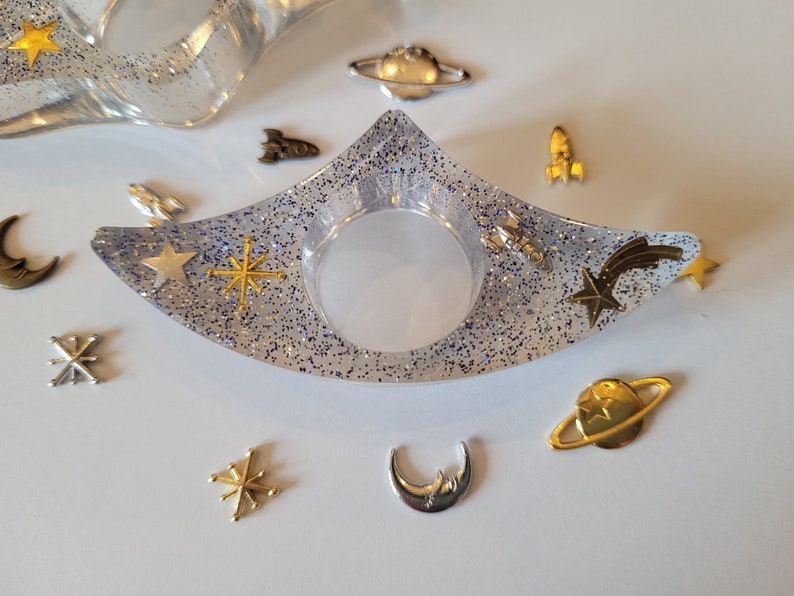 K&ouml;nnte beinhalten: Eine klare, mit Glitzer gef&uuml;llte Ring Schale mit einem Halbmond, Sternen und einem Kometen Design. Die Schale ist umgeben von kleinen goldenen und silbernen Charms, darunter Planeten, Sterne und Raketen.