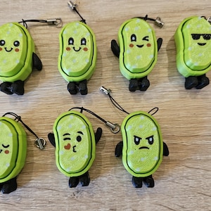 May include: Seven green bean-shaped charms with different facial expressions. Each charm has black arms and legs, and a black cord with a silver clasp. The charms are on a light brown wooden surface.