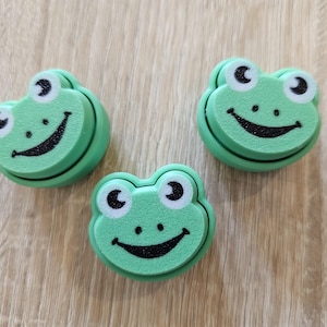 May include: Three green frog-shaped decorations with white eyes and a black smiling mouth are arranged on a light brown wooden surface. The frog's body is a light green colour.