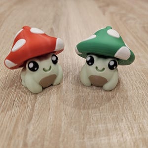 May include: Two charming mushroom frog figurines. One has a red mushroom cap with white spots, the other a green cap with white spots. Both frogs have light green bodies, brown bellies, and smiling faces.