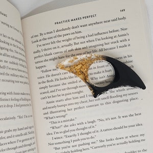 May include: A close-up of a book open to a page with the text "Practice Makes Perfect" and a black bookmark with gold accents. The text on the page discusses the importance of practice and the impact of a bad influence. The bookmark is shaped like a sharp, pointed object.
