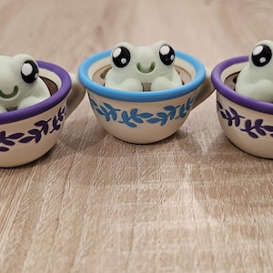 May include: Three ceramic frog figurines with large black eyes and smiling mouths sit inside decorative teacups. The teacups have a white base with purple and blue accents and leaf patterns. The teacups are on a wooden surface.