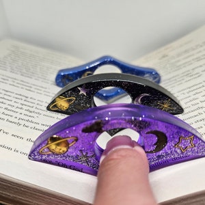 K&ouml;nnte beinhalten: Drei &uuml;bereinander gestapelte Lesezeichenringe aus Harz mit Galaxie-Motiv. Der obere Ring ist lila mit goldenem Glitzer, einer Sichelmond, einem Stern und einem Planeten mit Ringen. Der mittlere Ring ist schwarz mit einem silbernen Stern und einer Sichelmond. Der untere Ring ist blau mit einem silbernen Stern und einer Sichelmond. Die Ringe sind so konzipiert, dass sie eine Seite in einem Buch festhalten.