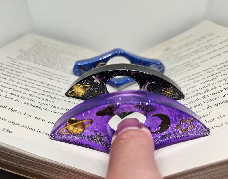K&ouml;nnte beinhalten: Drei &uuml;bereinander gestapelte Lesezeichenringe aus Harz mit Galaxie-Motiv. Der obere Ring ist lila mit goldenem Glitzer, einer Sichelmond, einem Stern und einem Planeten mit Ringen. Der mittlere Ring ist schwarz mit einem silbernen Stern und einer Sichelmond. Der untere Ring ist blau mit einem silbernen Stern und einer Sichelmond. Die Ringe sind so konzipiert, dass sie eine Seite in einem Buch festhalten.