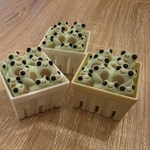 May include: Three small, square baskets filled with light green frog-shaped sweets. The frogs have black eyes and brown bellies. The baskets are white and beige, on a wooden surface.