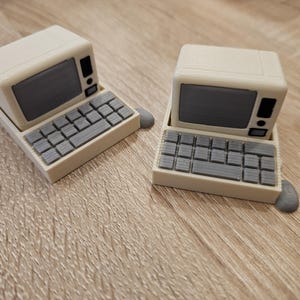 May include: Two miniature retro computer models. Each has a cream body, a grey screen, and a grey keyboard. The computers are placed on a wooden surface. The models are small, with a mouse attached to each.