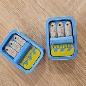 May include: Two blue sardine cans, each containing three fish. The cans feature a yellow and blue label with the word "SARDINES". The fish are white with black eyes. The lids are partially open, revealing the fish inside.